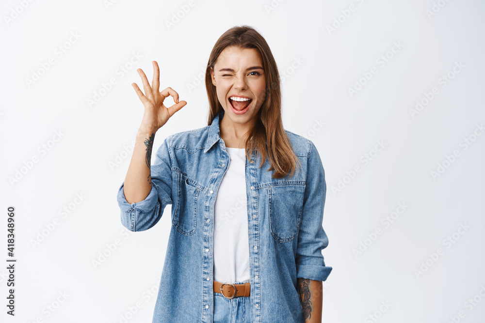 Cheerful young woman winking and showing OK sign, say alright or yes, approve something good, recommending product and quality, standing pleased against white background Cheerful young woman winking and showing OK sign, say alright or yes, approve something good, recommending product and quality, standing pleased against white background
