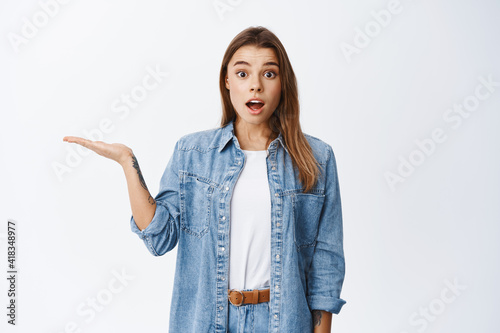 Portrait of surprised beautiful girl holding in hand, spread hand sideways and showing object in palm and smiling amazed, standing on white background