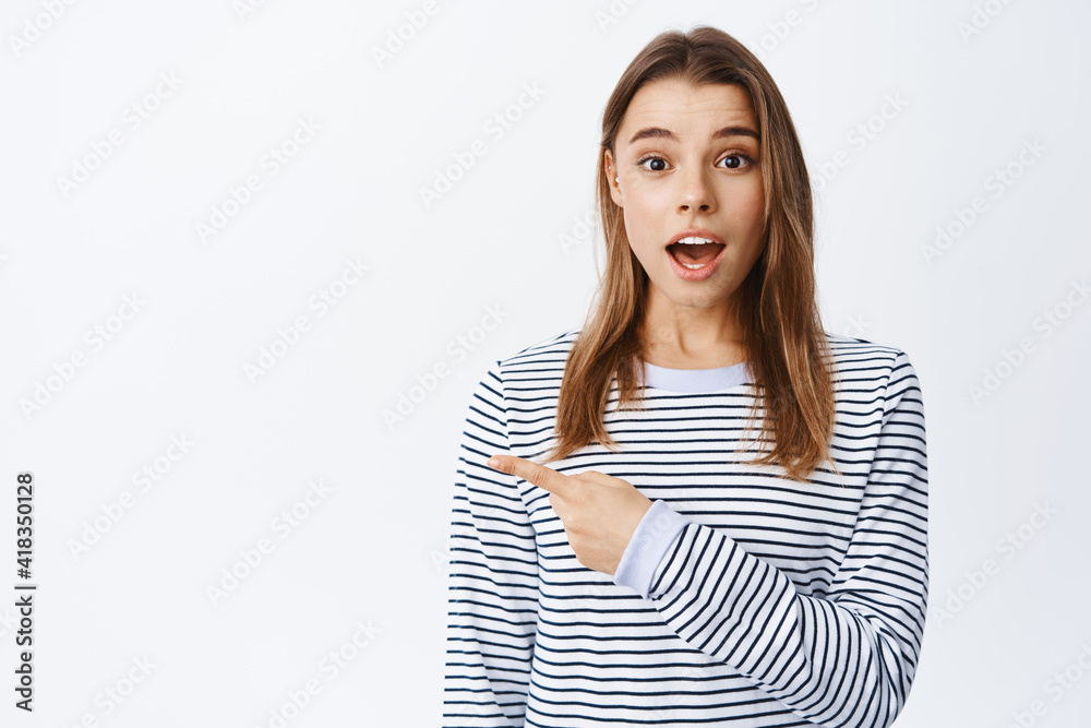 Surprised young woman with blond hair pointing left at logo, open mouth fascinated, checking out promo offer, standing over white background Surprised young woman with blond hair pointing left at logo, open mouth fascinated, checking out promo offer, standing over white background