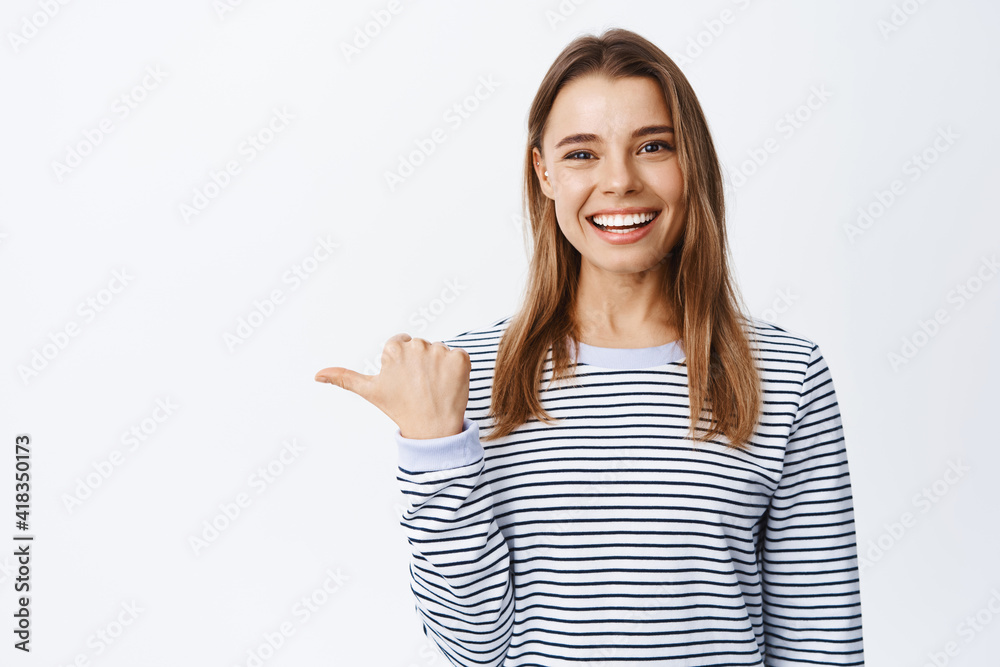 Portrait of cheerful female model with white teeth pointing left, inviting to check out or look at logo, showing advertisement, white background Portrait of cheerful female model with white teeth pointing left, inviting to check out or look at logo, showing advertisement, white background