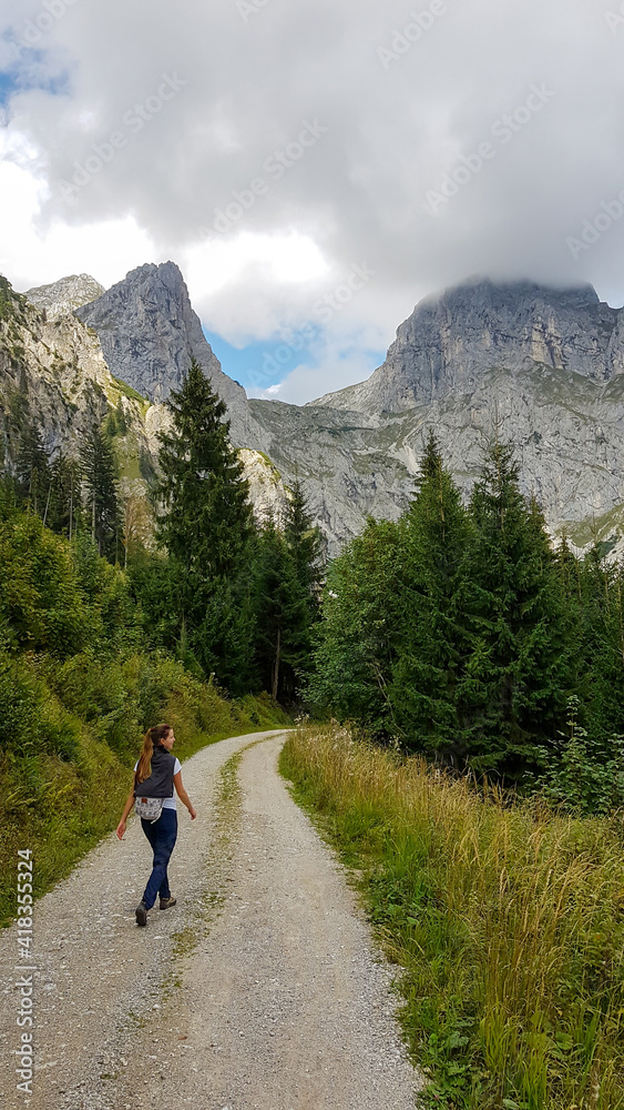 A backpacker woman hiking through Austrian Alps to the top of Hochkogel. She is walking on a gravelled pathway through a forest. High and sharp mountains are shrouded with thick fog. Outdoor activity