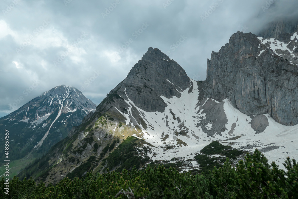 A panoramic view on the Alpine peaks in Austria from Marstein. The slopes are mostly covered with snow. Stony and sharp mountains. Overcast. Baren slopes, green valley below. Serenity and calmness