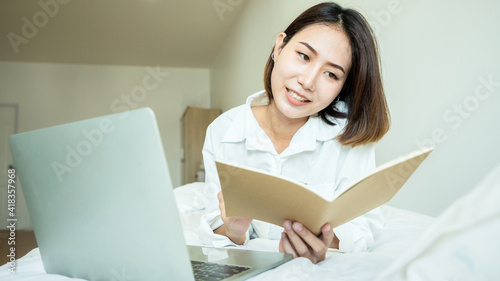 Happy casual beautiful Asian woman working on a laptop with book and laying on the bed. Working from home in quarantine lockdown.