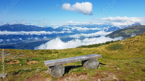 A wooden bench with a panoramic view on the Alpine valley from the top of Granattor in Austrian Alps. The valley is shrouded in fog. The slopes are lush green. Many mountain poking above the clouds photo