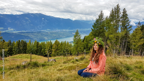 A woman in orange hoodie wearing a floral crown made of wild flowers and larch branches. Lush green pasture and dense forest. Many mountains poking above the clouds. Millstaettersee lake in the back. photo