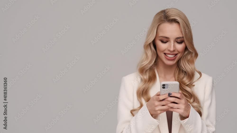 A smiling young blonde businesswoman is using her smartphone standing in the side isolated over gray background in studio A smiling young blonde businesswoman is using her smartphone standing in the side isolated over gray background in studio