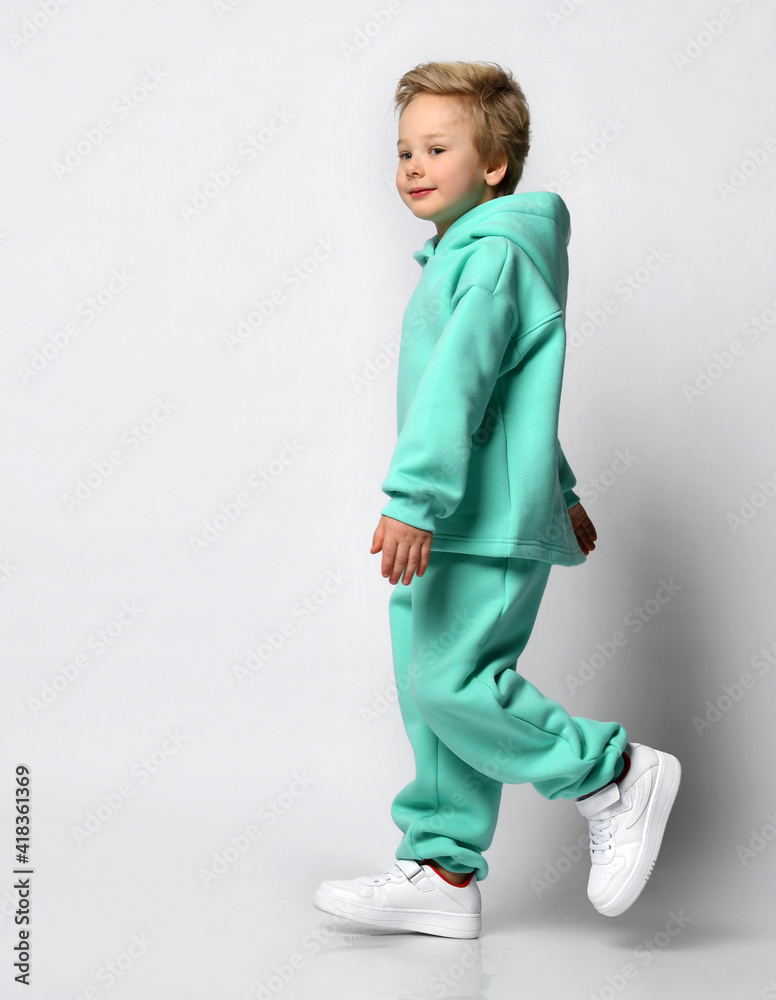 Full length portrait of a little stylish boy in a warm sports suit on a gray background. Boy dressed in a turquoise fleece suit and sneakers stands sideways to the camera near a free space for text. Full length portrait of a little stylish boy in a warm sports suit on a gray background. Boy dressed in a turquoise fleece suit and sneakers stands sideways to the camera near a free space for text.