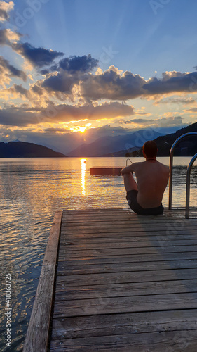 A man in full cup sitting at the end of a wooden pier of Millstaetter lake and enjoys the sunset. The sun sets behind high Alps. Calm surface of the lake reflects the orange sky and the mountains photo