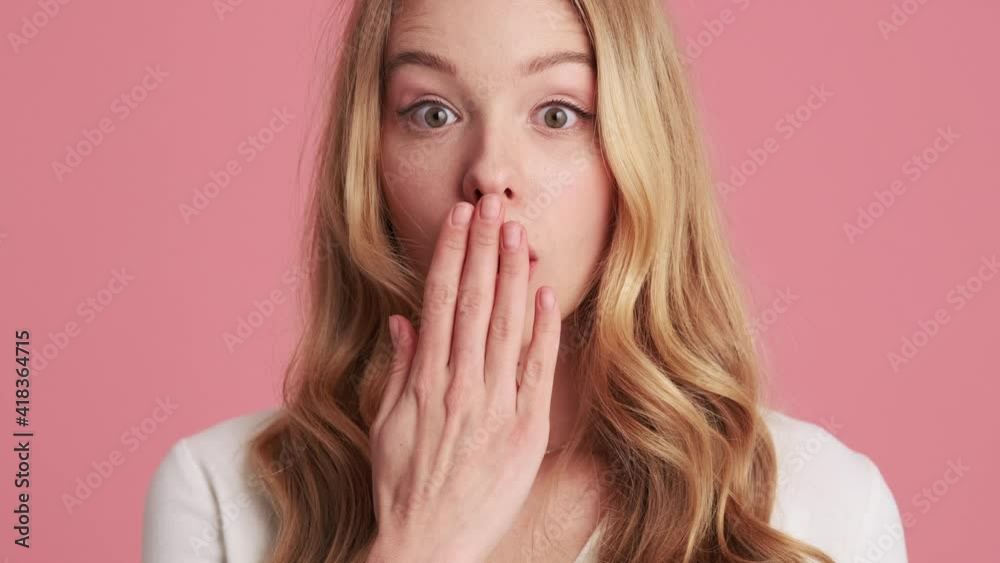 A charming young blonde woman is looking to the camera while covering her mouth with her hands standing isolated over a pink wall in the studio A charming young blonde woman is looking to the camera while covering her mouth with her hands standing isolated over a pink wall in the studio