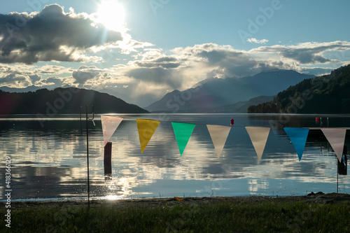 A sunset by Millstaetter lake in Austria. The lake is surrounded by high Alps. Calm surface of the lake reflecting the sunbeams. A row of colorful flags hanging at the shore. A bit of overcast. Calm photo