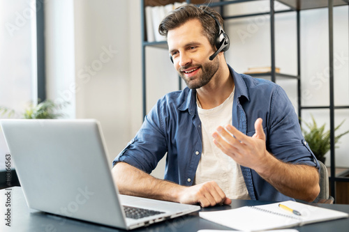 Confident man with handsome stubble, wearing headset, talking in front of computer, wearing casual clothes, talking at computer, discussing work process, gesticulates, chatting with partners, notice