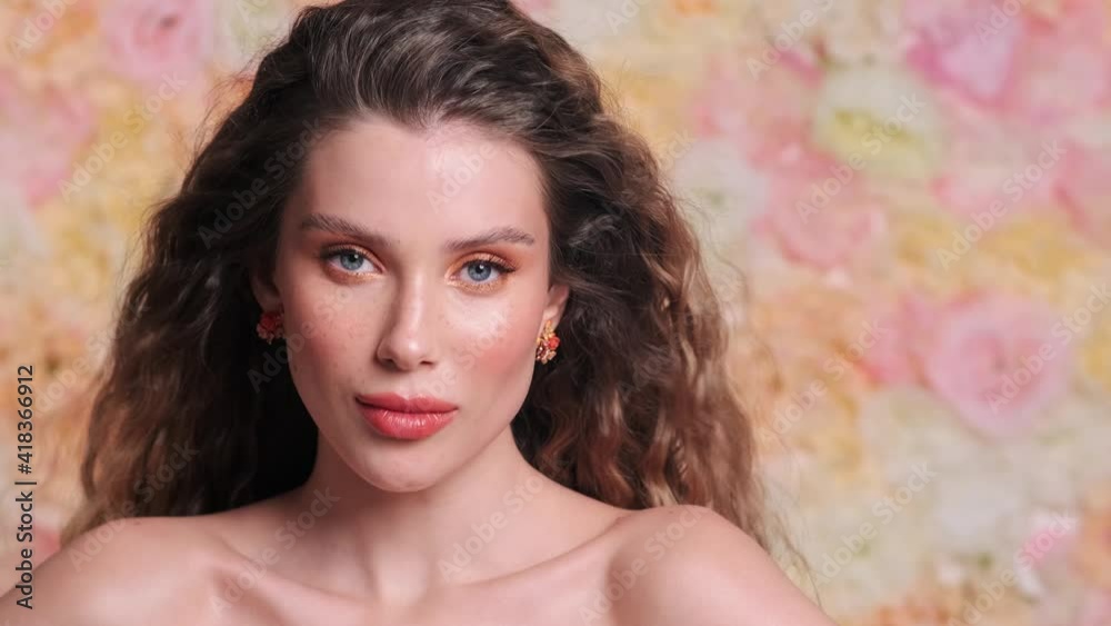 A close-up view of a beautiful young woman is posing to the camera while hair fluttering in the wind standing isolated over a floral background in the studio A close-up view of a beautiful young woman is posing to the camera while hair fluttering in the wind standing isolated over a floral background in the studio