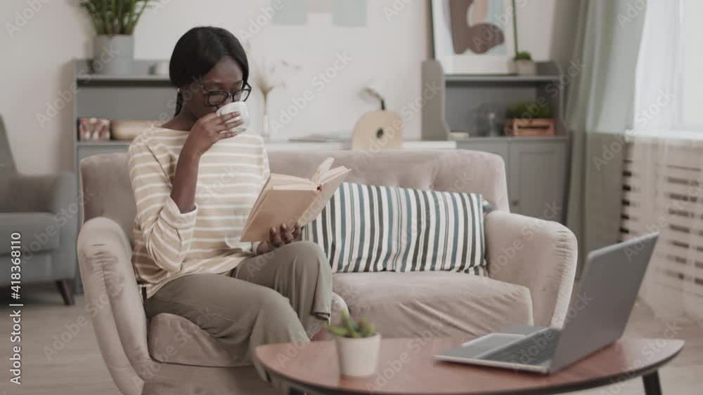 Lockdown of young African-American woman wearing casual clothes and glasses sitting on couch in living room, reading book and drinking coffee Lockdown of young African-American woman wearing casual clothes and glasses sitting on couch in living room, reading book and drinking coffee