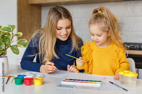 Mother and daughter painting at home. Cute little kid in yellow sweater having fun with parent and paints. Concept of early childhood education, hobby, talent, preschool leisure and parenting