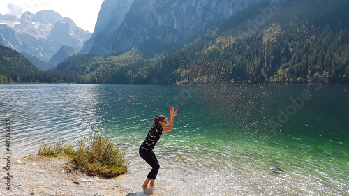 A woman squatting by the shore of Gosau lake and sprinkling the water with her arms. The lake is surrounded by high mountains, with Dachstein glacier in the back. The woman is having fun. Happy moment
