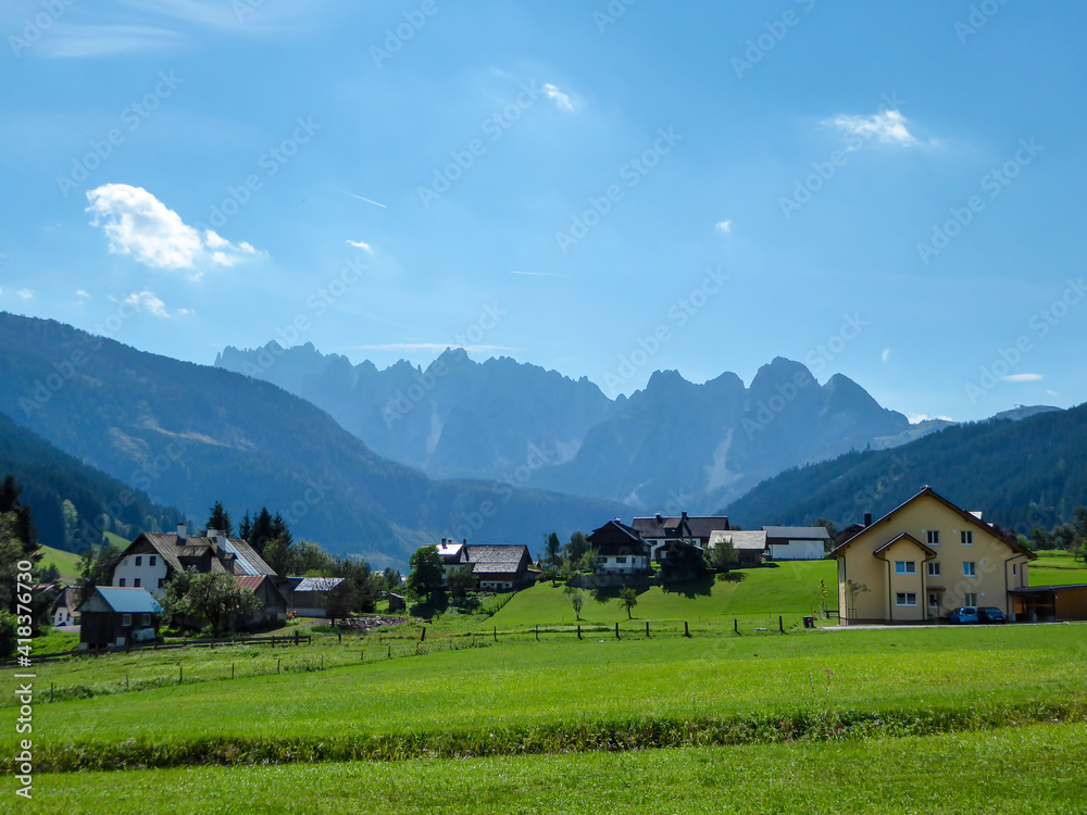 A panoramic view on the distant, high Alpine chains in the region of Gosau, Austria. There is a small village along the lush pasture in front. There is a little bit of a haze around. Sunny day.