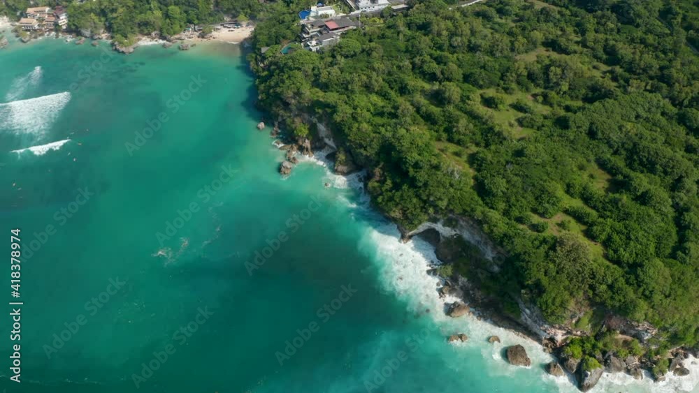 Aerial view of waves crashing into rocky cliff on the Bali coastline. Luxury resorts on lush tropical coastline by a turquoise blue sea Aerial view of waves crashing into rocky cliff on the Bali coastline. Luxury resorts on lush tropical coastline by a turquoise blue sea