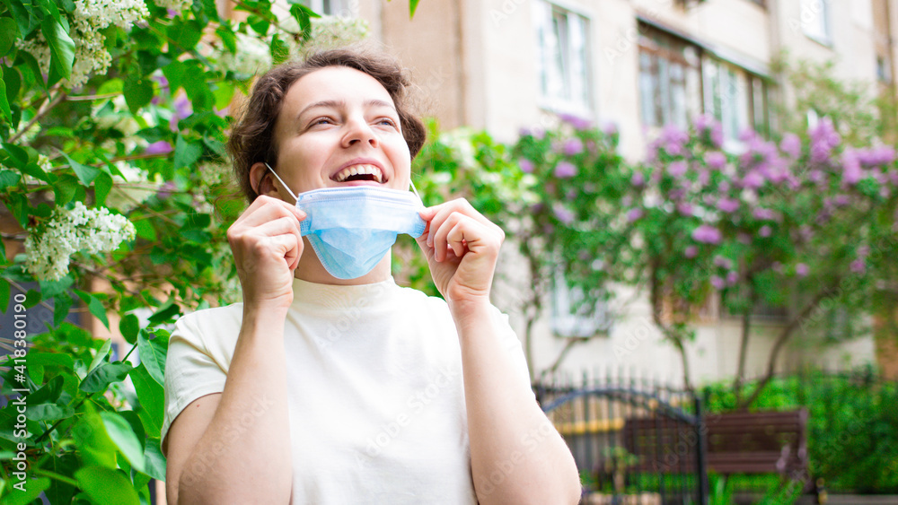 Smiling happy girl taking off medical protective mask outdoor, breathing deep with pleasure, standing on blooming lilac trees and house background outdoors. Spring allergy. Freedom. End of quarantine Smiling happy girl taking off medical protective mask outdoor, breathing deep with pleasure, standing on blooming lilac trees and house background outdoors. Spring allergy. Freedom. End of quarantine