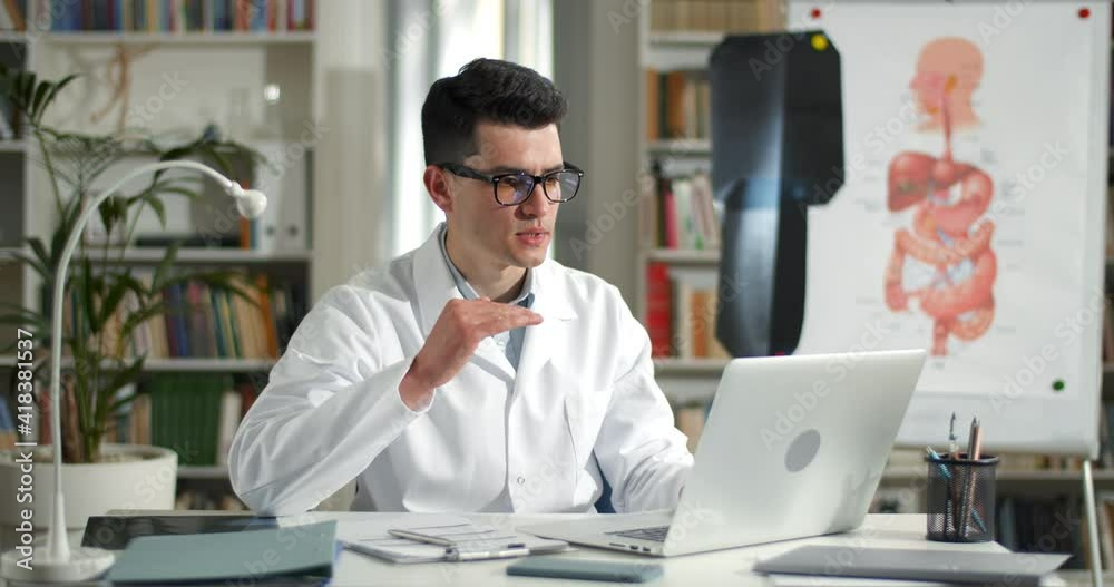 Serious man in glasses and professional white gown talking and gesturing while having online consultation. Male doctor looking to laptop screen while sitting at table in medical office Serious man in glasses and professional white gown talking and gesturing while having online consultation. Male doctor looking to laptop screen while sitting at table in medical office