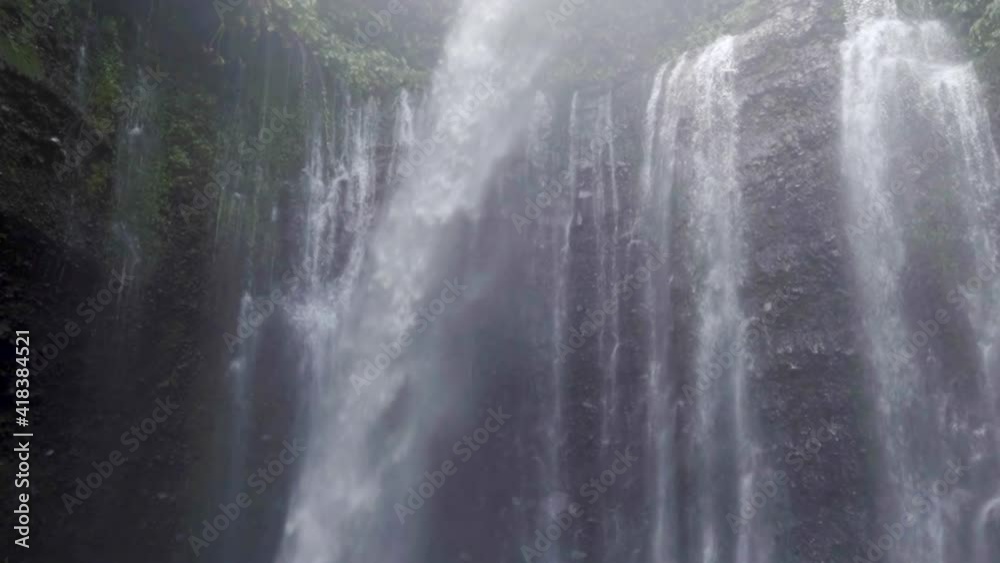 Tilt down along the Sendang Gile Waterfalls with mist, vine covered ledge walls, rocks, and shirtless man - Nusa Penida & Lombok, Bali Tilt down along the Sendang Gile Waterfalls with mist, vine covered ledge walls, rocks, and shirtless man - Nusa Penida & Lombok, Bali