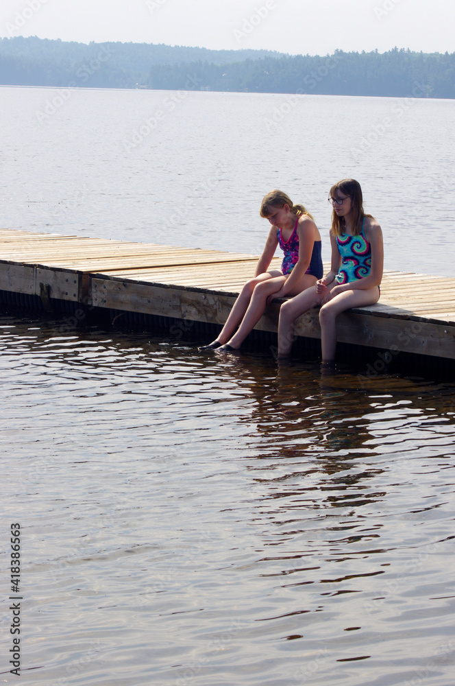 two young girls sitting in swimsuits on wooden dock in water in summer with copy space