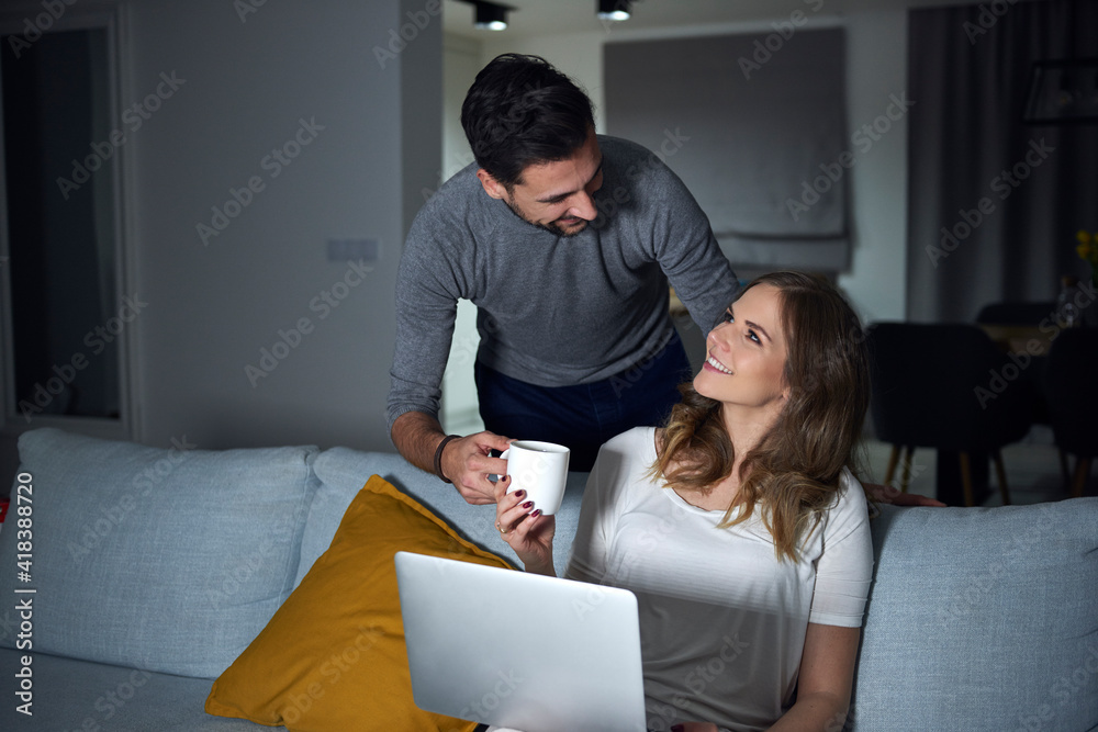 Young man handing coffee to girlfriend sitting on sofa in evening Young man handing coffee to girlfriend sitting on sofa in evening