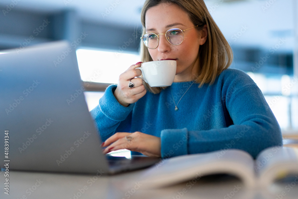Female higher education student looking at laptop and drinking coffee in university cafe, close up Female higher education student looking at laptop and drinking coffee in university cafe, close up