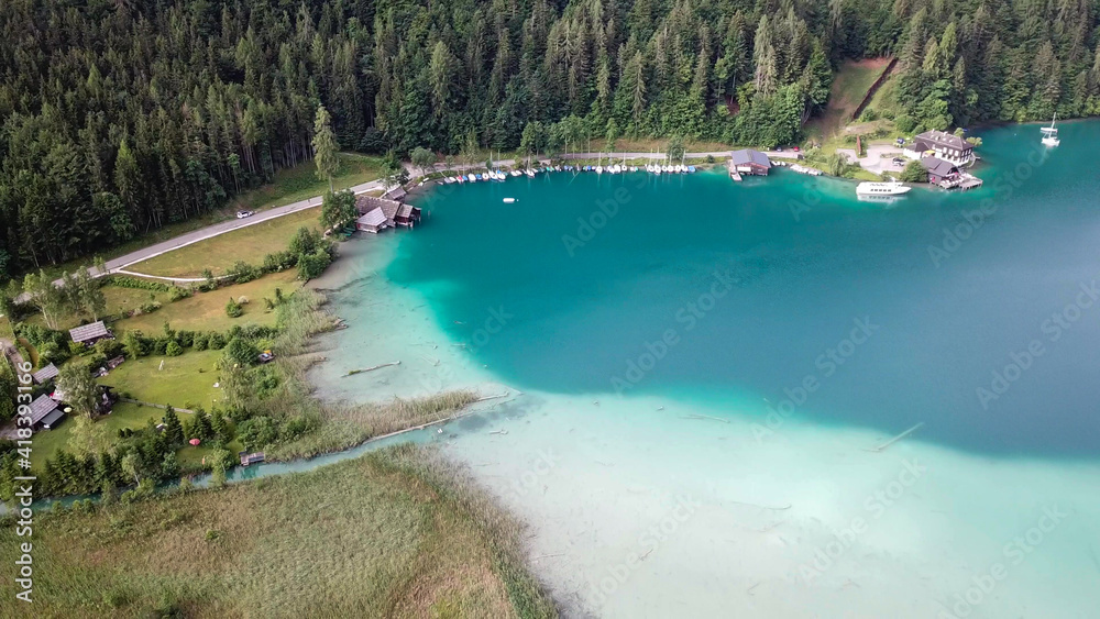 A top down, drone capture of Weissensee lake in Austrian Alps. The water changes color from white, turquoise to navy blue. A small settlement at the shore. A bit of overcast. Serenity and peacefulness