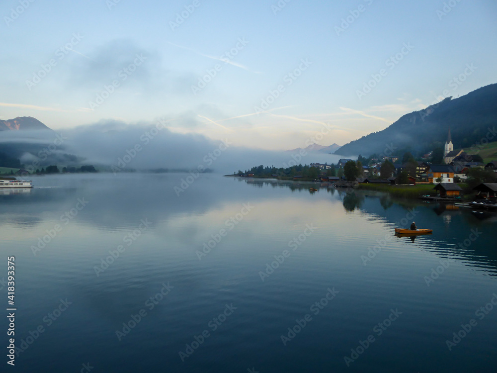 An idyllic, panoramic view on the Weissensee lake in Austria. The lake is surrounded by high Alps. There is a small village at the lake's shore, with tall church tower. Few clouds. Calmness and peace