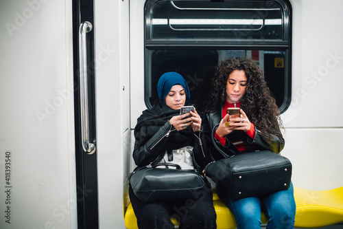 Young woman in hijab and friend sitting on subway train looking at smartphones
