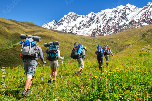 A group of tourists with large backpacks climbs the grassy slope overlooking the snow-capped peaks in the Caucasus Mountains Georgia Svaneti