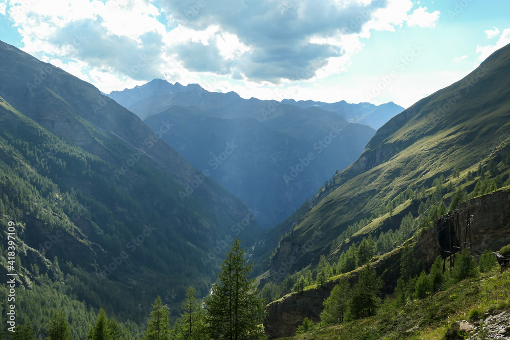A panoramic view on vast valley with the view on Grossglockner in Heiligenblut region in Austria. The  valley has lush green color. There are high Alpine chains in the back. A bit of overcast