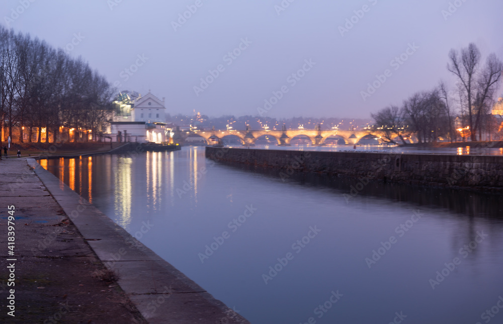 Vltava river bank after sunset and in the background blurred Charles Bridge in Prague