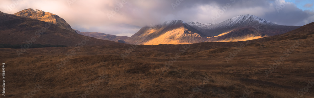 A wide panorama view of dramatic dappled light over a mountain countryside landscape near Glencoe in the Scottish Highlands, Scotland. A wide panorama view of dramatic dappled light over a mountain countryside landscape near Glencoe in the Scottish Highlands, Scotland.
