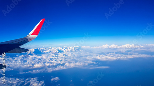 Window view from the plane on snow capped Himalayan ranges in Nepal. The high peak are shrouded in clouds. Lots of clouds below. Wing of the plane in the corner. Flying high above the ground. Freedom