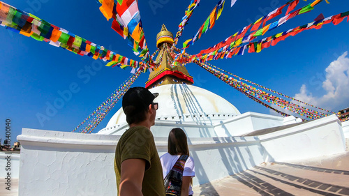 A couple walking along the prayer flags hanging from the top of boudhanath Temple in Kathmandu, Nepal. The flags have 'om mani padme hum' mantra written on them. The round temple is painted white. photo