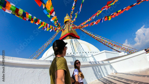 A couple walking along the prayer flags hanging from the top of boudhanath Temple in Kathmandu, Nepal. The flags have 'om mani padme hum' mantra written on them. The round temple is painted white. © Chris
