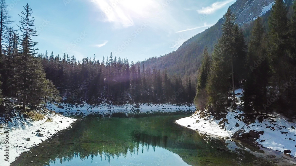 A drone shot of Green Lake in Austrian Alps. The lake shimmers with many shades of green and turquoise. Thick forest around it. Winter in the mountains. There is snow on the ground. Wandering