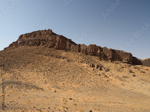 Ancient fortress on the top of the hill surrounded by the desert sand. Grey stones and grey ancient wall on the yellow sand, clear blue sky. 