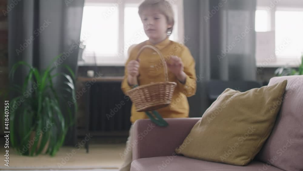 Medium shot of cute boy finding Easter eggs on couch at home and putting them in his basket