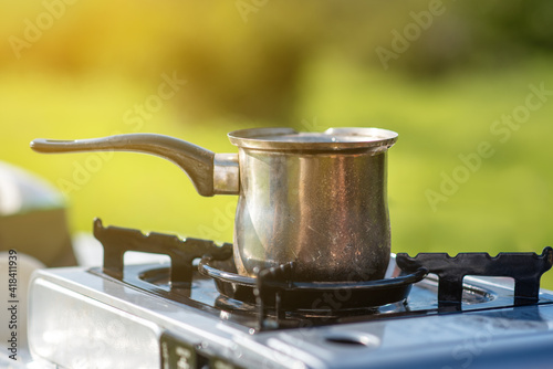 Making tea and coffee with a portable gas cooker on nature green background in the tourist camp on sunny day. Camping stove with small coffee pot in outdoors.
