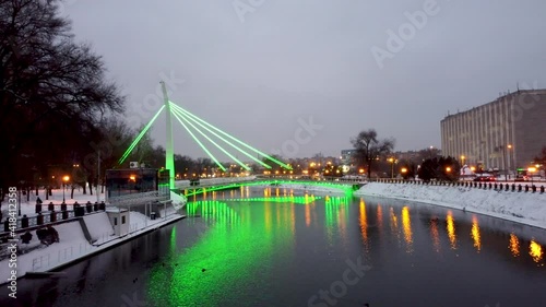 Pedestrian bridge (Mist Zakokhanykh) across river with green illumination reflected in dark water, Skver Strilka in Kharkiv city center. Winter aerial evening footage photo