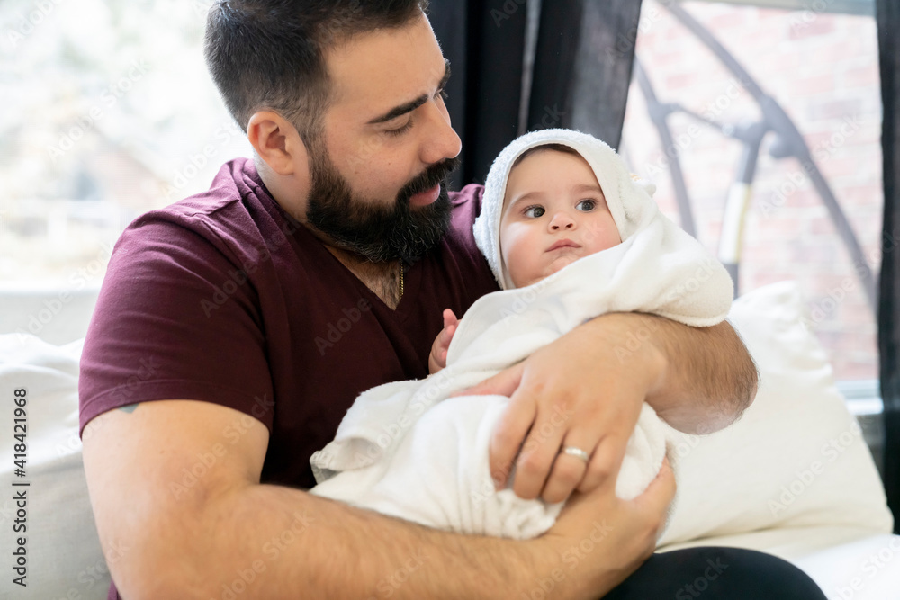 Man with beard laying on couch hugging chubby baby in white towel Man with beard laying on couch hugging chubby baby in white towel