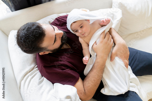 Man with beard laying on couch hugging chubby baby in white towel 