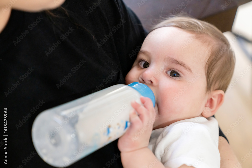 Woman feeding baby boy formula in a bottle in dining room of home Woman feeding baby boy formula in a bottle in dining room of home