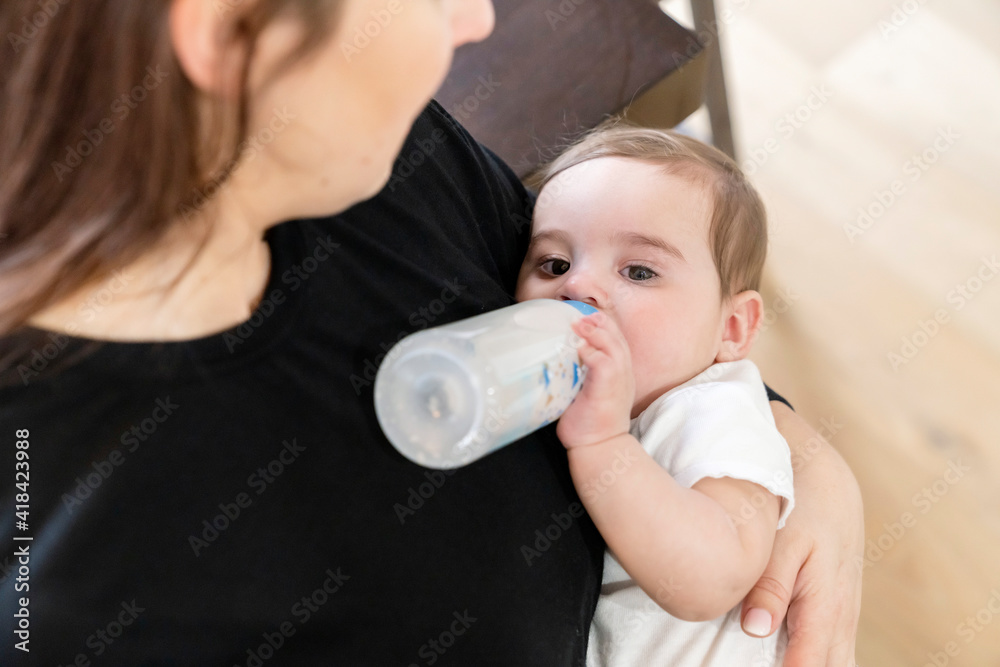 Woman feeding baby boy formula in a bottle in dining room of home Woman feeding baby boy formula in a bottle in dining room of home
