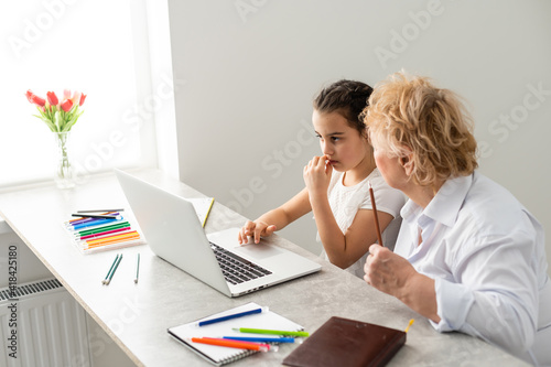 Woman tutor or foster parent mum helping cute caucasian school child girl doing homework sitting at table. Diverse nanny and kid learning writing in notebook studying at home.