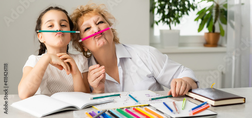 Woman tutor or foster parent mum helping cute caucasian school child girl doing homework sitting at table. Diverse nanny and kid learning writing in notebook studying at home.