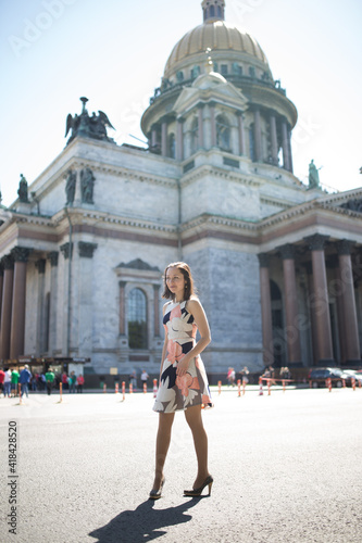 A very beautiful brunette woman in heels and in a dress walks lightly along the cathedral. She smiles and looks into the camera.