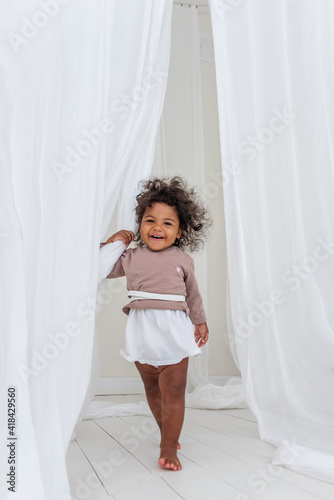 Little curly African American girl has fun laughs dances among airy white tulles fabric. Minimalist interior, Scandinavian style. Happy childhood. The concept of adopted children. Mixed ethnicity race © farmuty